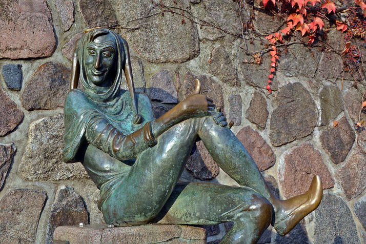 A bronze statue of a whimsical figure in a jester's outfit, seated with a playful expression, giving a thumbs-up gesture, set against a stone wall adorned with trailing red ivy.