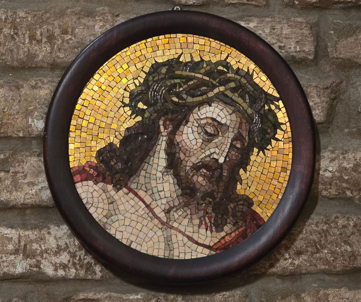A circular mosaic artwork depicting Jesus Christ wearing a crown of thorns. The mosaic features golden background tiles, a wooden frame, and is mounted on a stone wall.