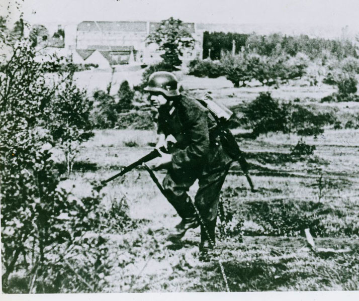 A German soldier in uniform and helmet crouches with a rifle in a rural area, likely during World War II combat operations.