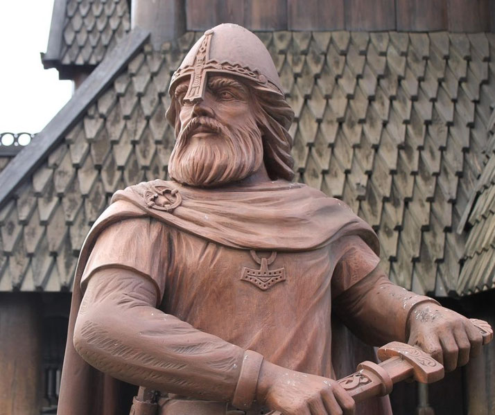 A wooden statue of a Viking warrior with a beard, helmet, and sword stands in front of a shingled wooden building.
