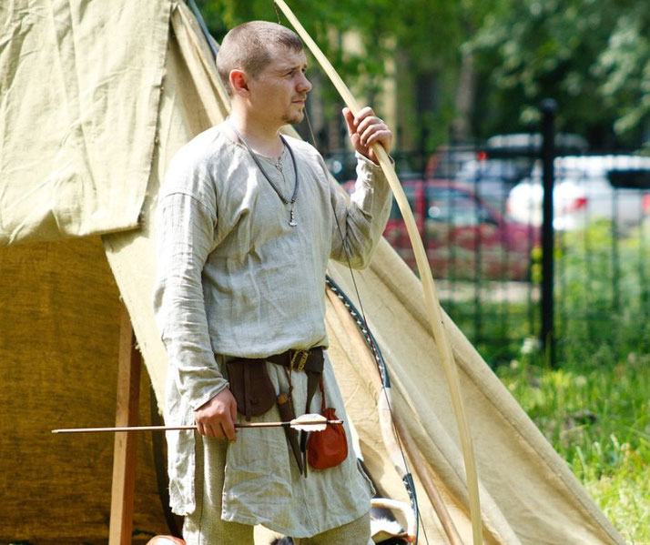 A man dressed in medieval-style linen clothing holds a longbow and an arrow, standing near a canvas tent.
