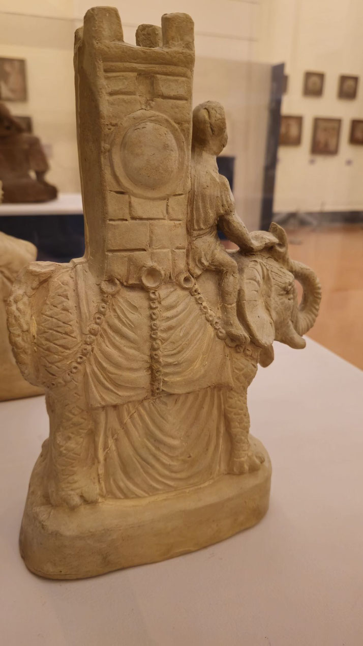 Ancient-style sculpture of a war elephant draped in armor, carrying a tower with a rider seated on its back.