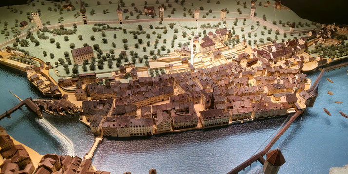 A detailed scale model of a historical riverside town with medieval architecture, bridges, watchtowers, and surrounding greenery, capturing a vibrant urban layout near a waterway.