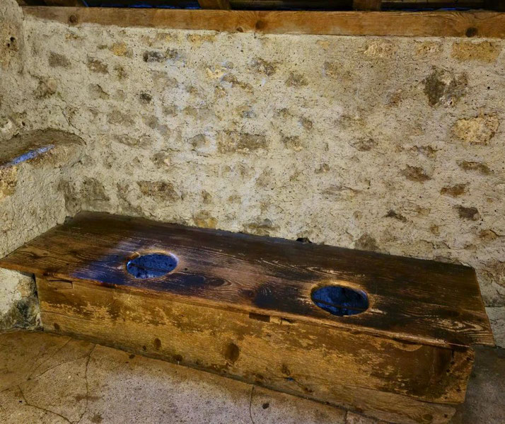 A medieval wooden toilet bench with two openings sits against a stone wall. The worn wood and rustic setting reflect historical sanitation practices in castles and communal hygiene of the time.