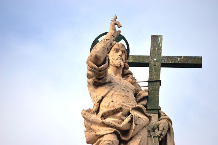 Stone statue of Jesus with a raised hand gesture, halo behind his head, and a large cross by his side against a pale sky.