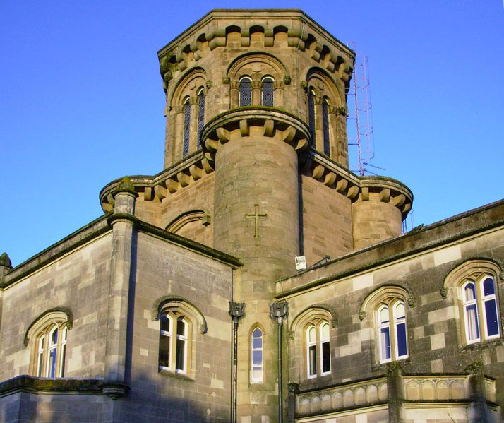 A view of a stone building with a rounded turret, featuring arched windows and a cross on the exterior.