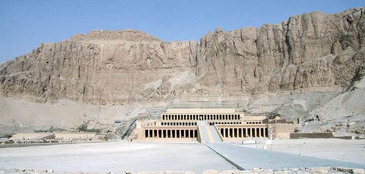 Ancient Egyptian temple with colonnaded terraces set against a massive rocky cliff under a clear sky.
