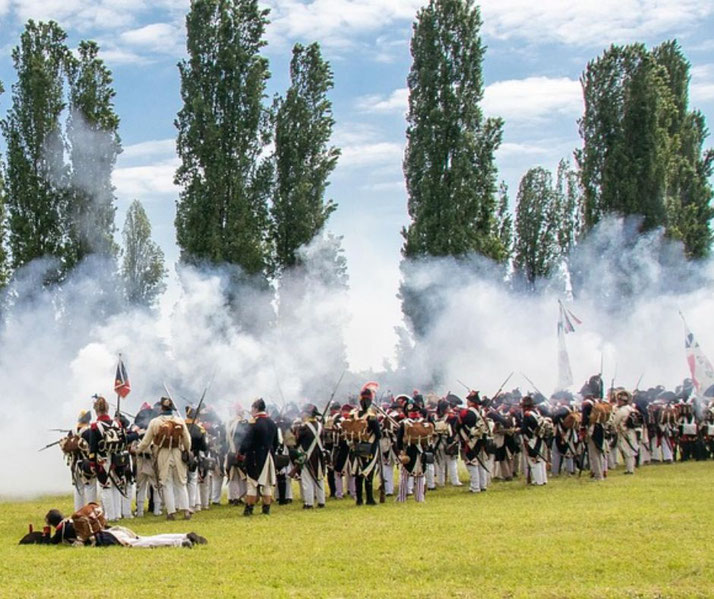 A Napoleonic-era battle reenactment, with soldiers in period uniforms fighting amid gun smoke on a grassy field.
