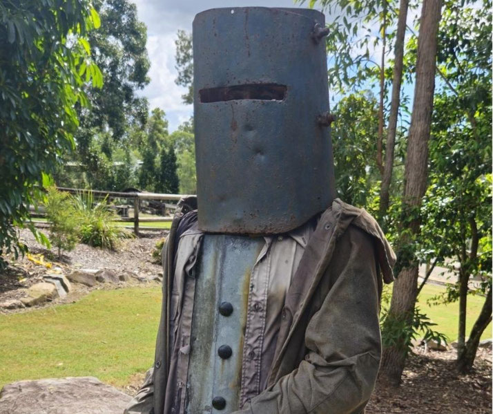 A person wears a large, rusted metal helmet with a narrow eye slit, evoking the appearance of Ned Kelly, in a leafy outdoor setting.