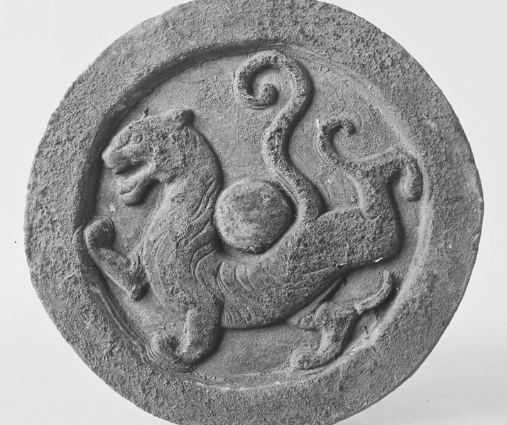 An ancient Chinese roof tile end with a carved tiger design, symbolizing strength and protection. The circular stone relief showcases intricate detailing and swirling patterns.