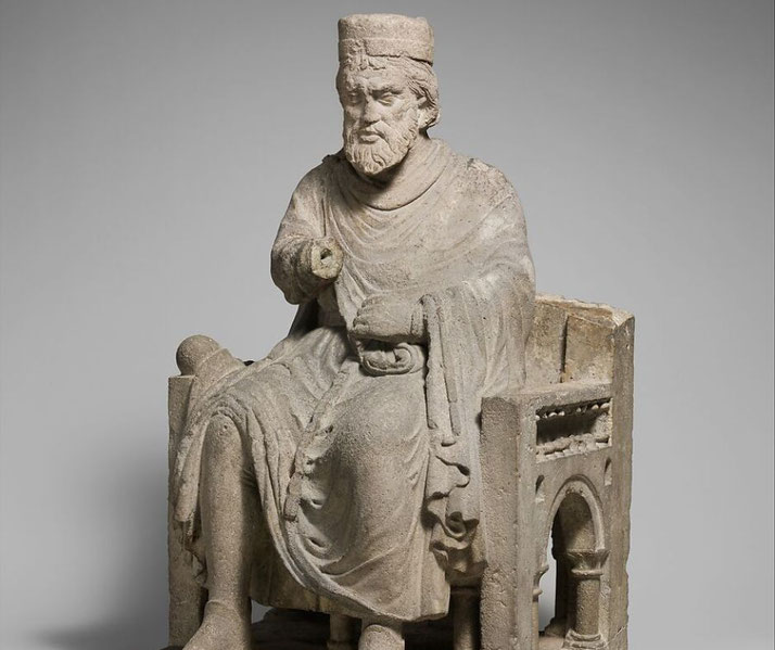 A weathered stone sculpture of a bearded king seated on an ornate throne. He wears a crown and flowing robes, holding an object in his right hand.