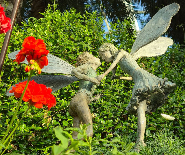 A garden statue of two fairies leans in for a kiss among lush greenery and red flowers.