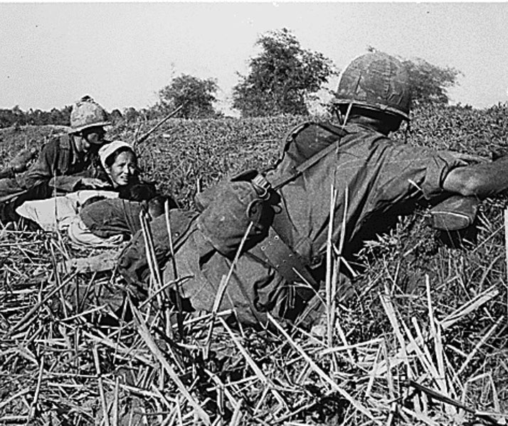 American soldiers lie prone in a field with a wounded Vietnamese civilian woman, providing aid and covering the area.