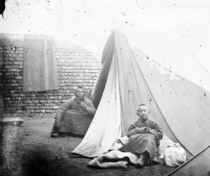 A black-and-white photo of two Mongolian individuals in front of a tent, with one sitting on the ground wrapped in blankets.