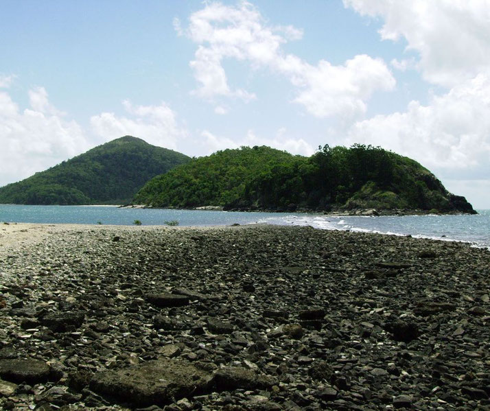 A rocky shoreline leading to a lush, green island surrounded by blue waters. The sky is partly cloudy, creating a serene and natural coastal landscape.