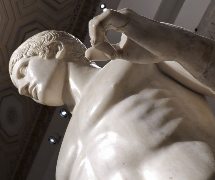Marble statue of a muscular male figure, viewed from below, with detailed hair and a raised hand forming a circle with thumb and finger.
