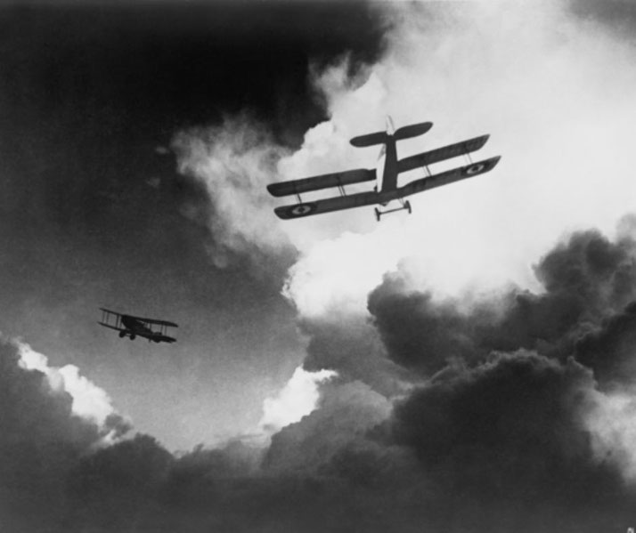 Two vintage biplanes with roundel markings fly through dramatic clouds in a black-and-white wartime sky scene.
