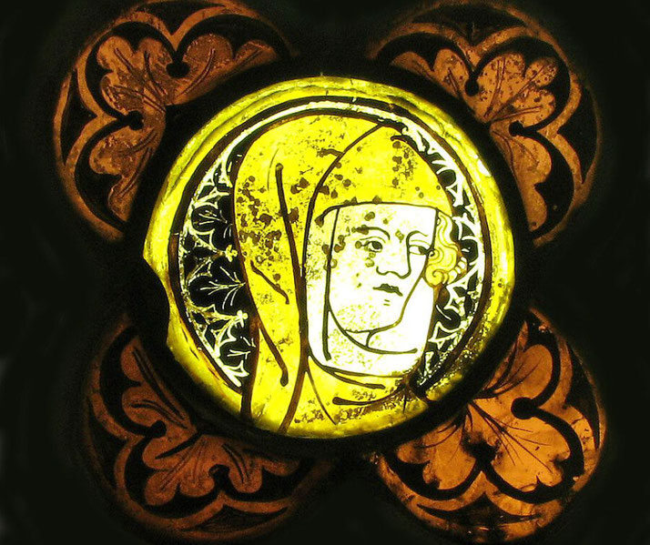 A medieval stained-glass window depicting a solemn-faced figure in a yellow hood, surrounded by intricate floral patterns in warm golden and black tones.