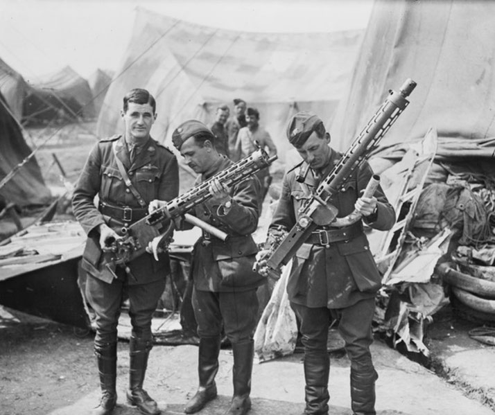 Three soldiers in uniform examine large aircraft machine guns near military tents, with others gathered in the background.