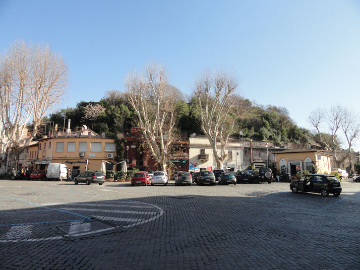 Small town square with parked cars, bare trees, and low-rise buildings at the base of a green, tree-covered hill under a clear blue sky.
