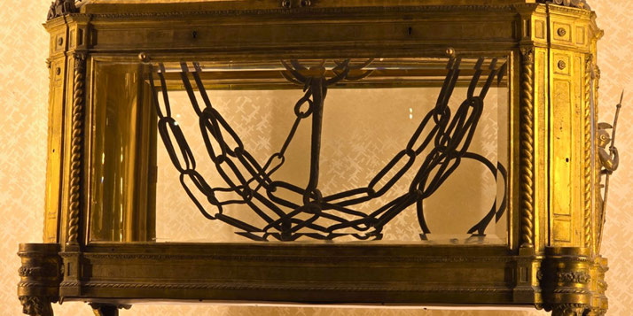 Ancient black chains displayed in a golden glass case, set against a patterned background, likely preserved as a religious or historical relic.