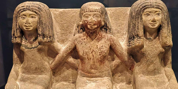 A limestone statue depicting an Egyptian man seated between two women, likely representing his wife and mother. The trio's hairstyles and attire reflect ancient Egyptian artistic conventions and familial ties.