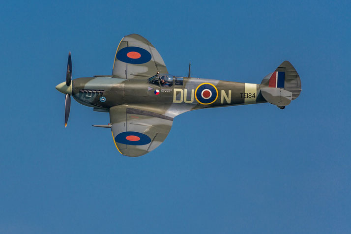 A Supermarine Spitfire with RAF markings flies against a clear blue sky, cockpit and roundels visible.