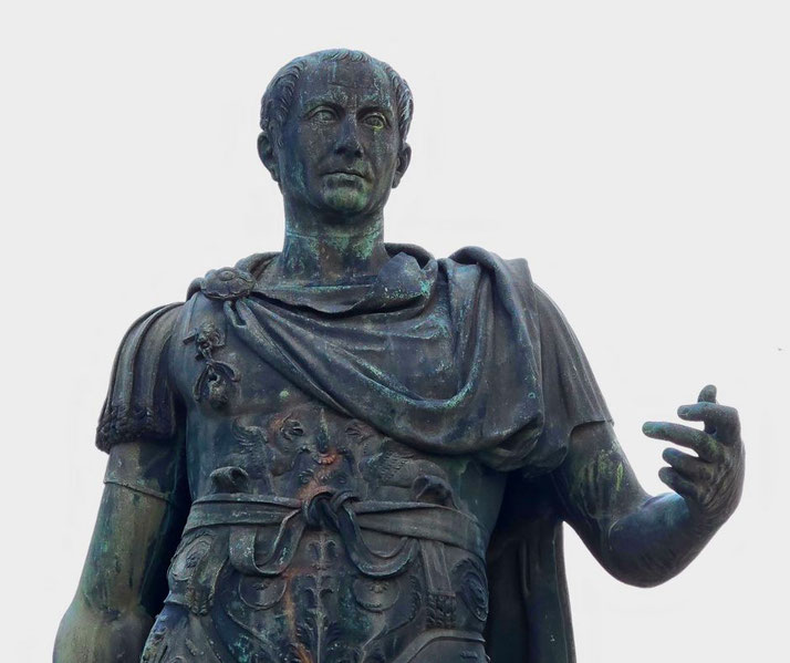 A bronze statue of a Roman leader in detailed armor and a draped cloak. He has a stern expression, cropped hair, and gestures with his right hand.