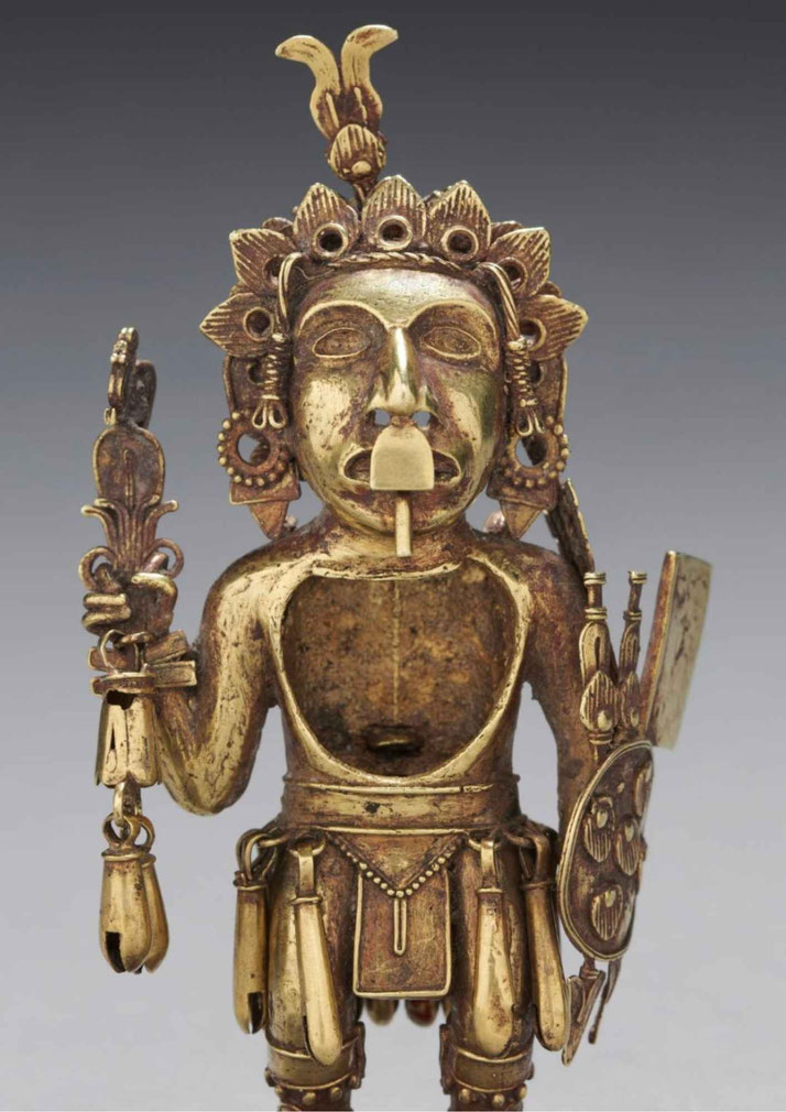 A gold figurine of a warrior, possibly from a Mesoamerican or South American culture. It features intricate details, including a headdress, shield, and ornaments, symbolizing status and power.