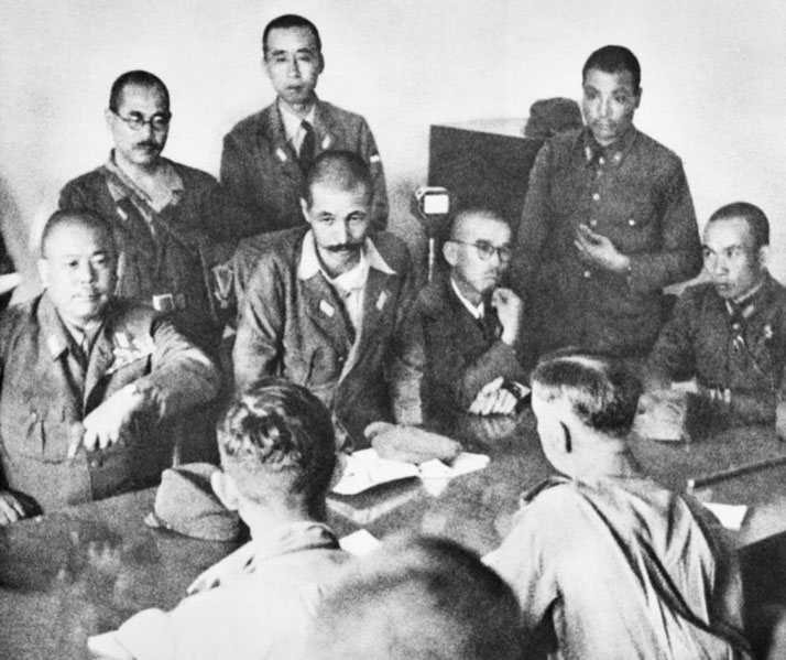 Military officers gather around a table during a wartime negotiation or surrender meeting in a formal setting.