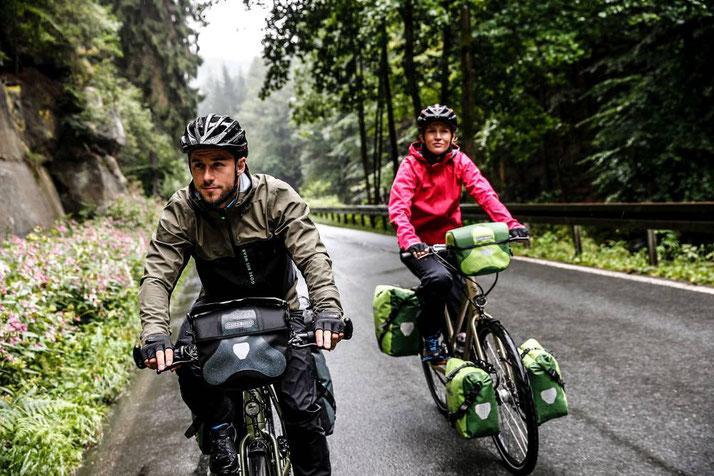 Pärchen fährt auf seinen e-Bikes mit den wasserfesten Taschen von Ortlieb über eine Waldstraße