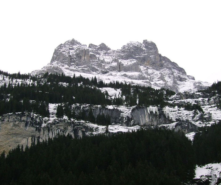 Snow-dusted rocky mountain rises above dense conifer forest, with exposed cliffs and overcast sky overhead.