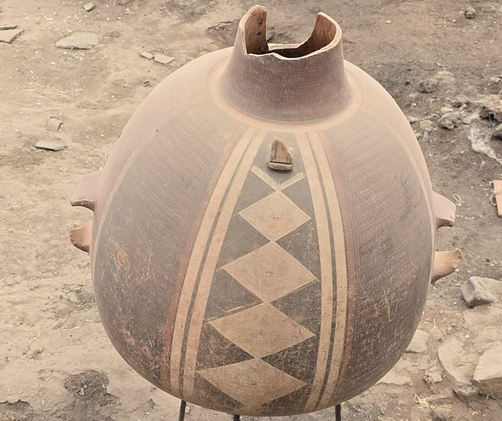 Large broken clay vessel with geometric diamond patterns, displayed on a stand in an archaeological dig site.