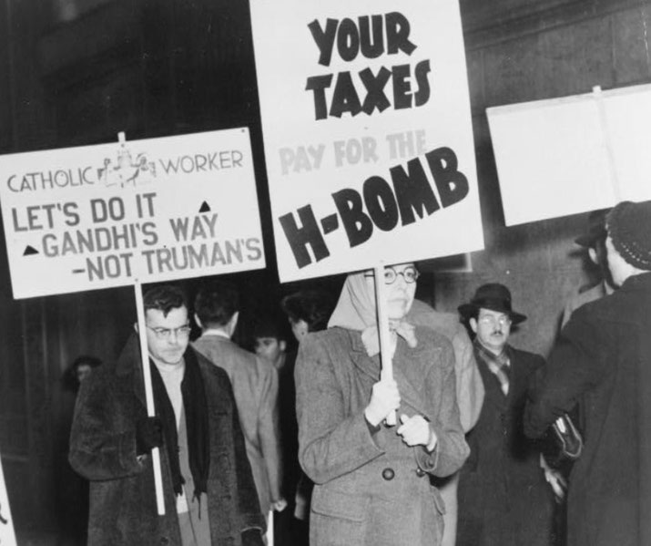 Black and white photo of protesters holding anti-nuclear signs, one referencing Gandhi and another criticizing tax funding for the H-bomb.