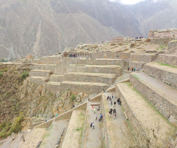Inca ruins of Ollantaytambo, Peru, with large stone terraces built into the mountainside. Tourists explore the site, while steep slopes and towering mountains form the backdrop.
