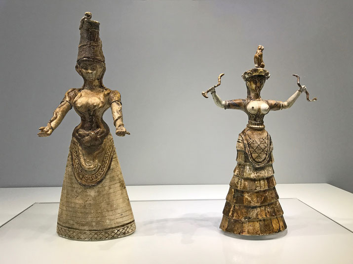 Two Minoan snake goddess figurines with raised arms and detailed attire displayed in a museum exhibit.