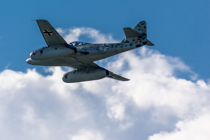 WWII-era German jet aircraft in flight against a backdrop of clouds and blue sky.