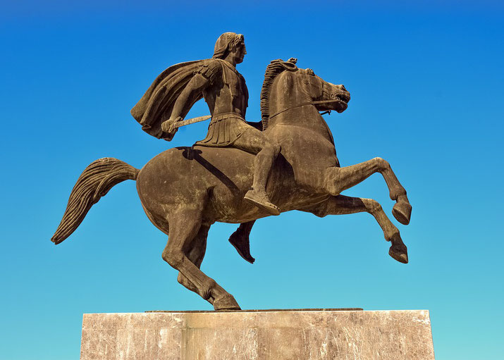 A bronze equestrian statue of Alexander the Great, with a flowing cape and sword, atop a rearing horse against a clear blue sky, symbolizing his military prowess and legacy.