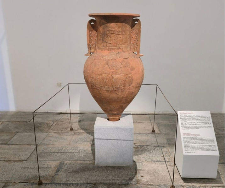 Ancient Mykonos Vase displayed on a museum pedestal, featuring terracotta relief scenes and large loop handles behind a low protective barrier.