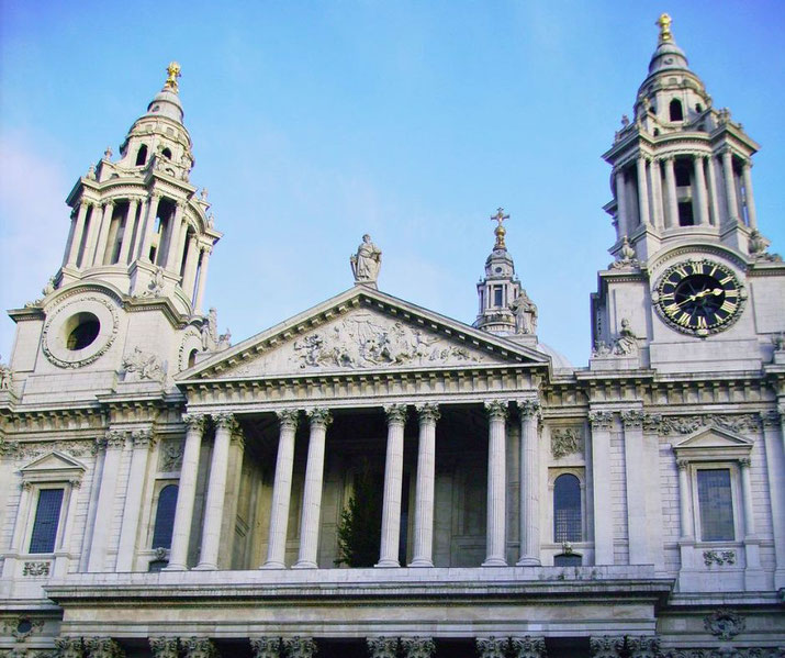A grand cathedral facade with twin bell towers, a clock face, tall columns, and statues under a clear blue sky.
