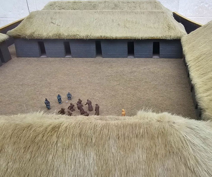 A detailed scale model of an ancient Inca courtyard features black stone walls, thatched roofs, and small figurines representing people engaged in activities.