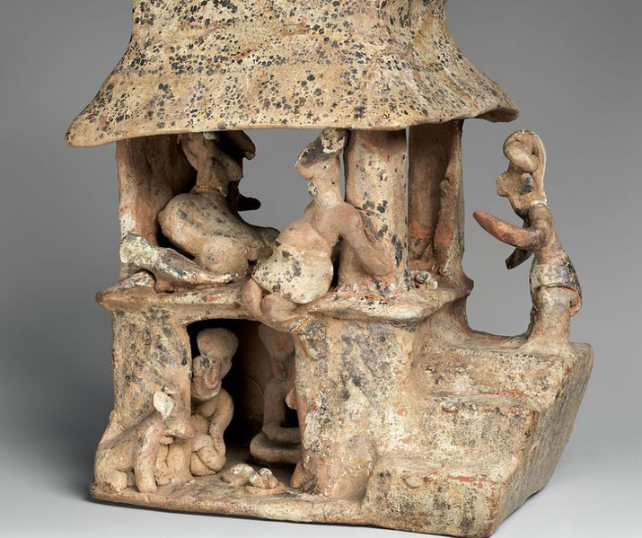 A terracotta sculpture of a two-story house with figures engaged in daily activities. The textured roof and worn details depict people sitting, interacting, and possibly preparing food, with animals present below.