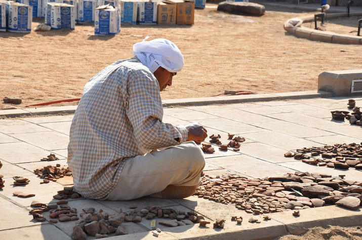 A man examines and organizes ancient pottery shards while sitting on the ground at an archaeological site.
