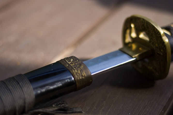 A close-up of a katana partially drawn from its black scabbard. The polished blade reflects light, and the ornate golden tsuba (handguard) features intricate detailing.