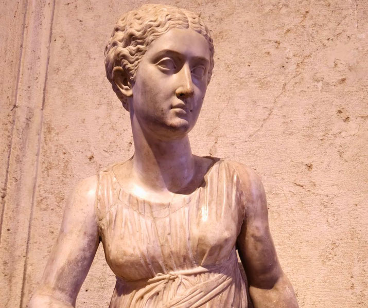 Marble statue of a woman with braided hair, wearing a belted, sleeveless gown, depicted in a graceful and serene pose against a textured stone background.