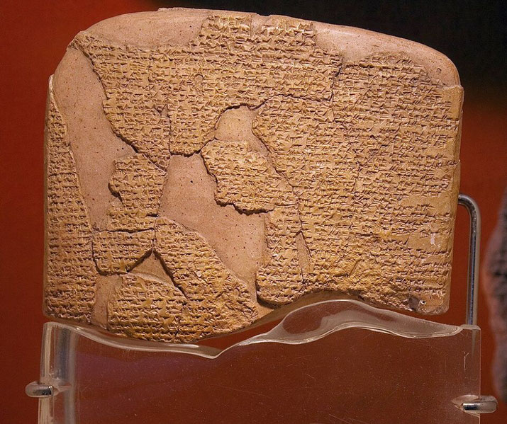 Ancient clay tablet inscribed with cuneiform script, partially broken, displayed on a clear stand against a reddish background.