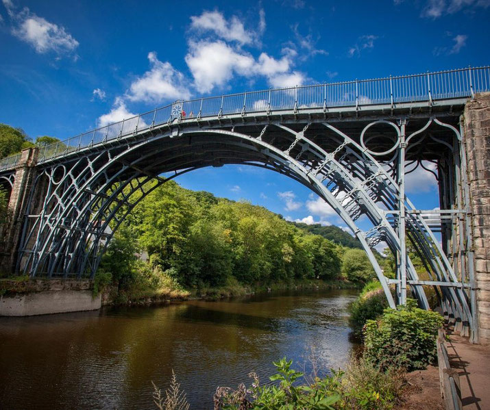 A historic iron bridge with intricate arches spans a calm river, surrounded by lush greenery under a bright blue sky with scattered clouds.
