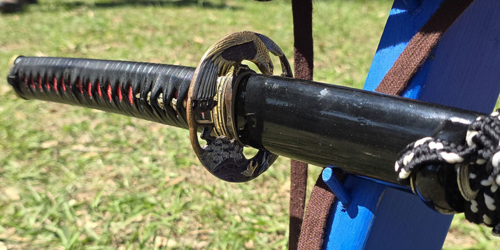 A close-up of a katana sword resting on a stand. The handle features intricate black and red wrapping, while the ornate handguard (tsuba) has a golden design.