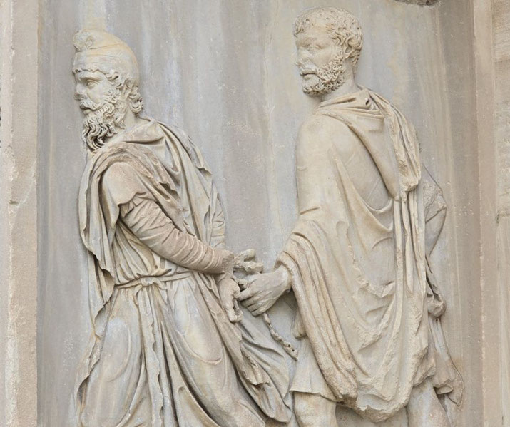 Stone relief of a Roman soldier holding a chained captive by the wrists, both figures in detailed robes, conveying a scene of conquest or arrest.