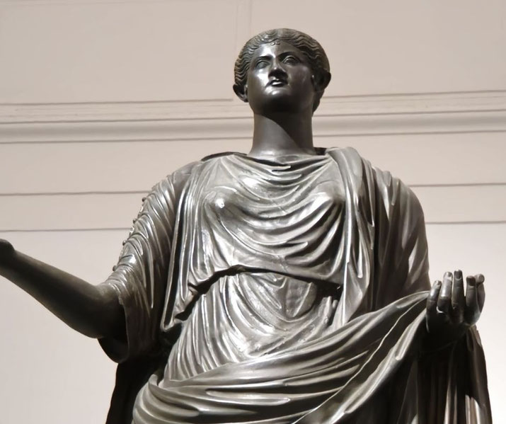 Bronze statue of a woman in flowing drapery, arms extended outward, with a serene, upward gaze and an elegant, poised stance.